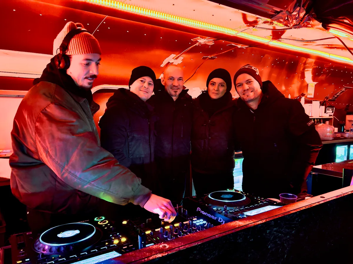 Man sieht ein DJ-Pult, an dem ein DJ gerade einen Regler betätigt. Hinter ihm halten sich drei Männer und eine Frau in den Armen und lächeln in die Kamera.